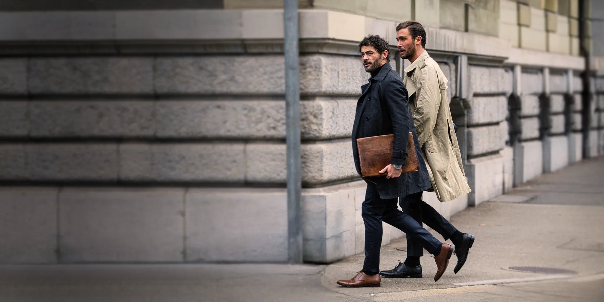 Two men in trench coats walking past a stone building